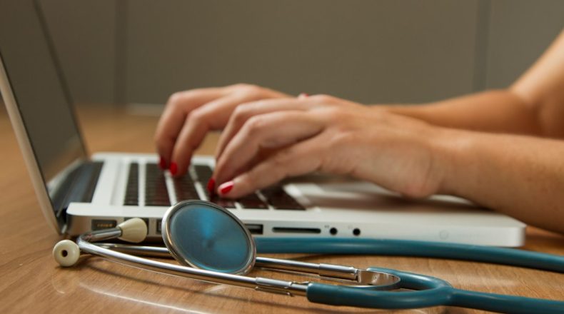 Close-up of hands typing on a laptop with a stethoscope in the foreground.